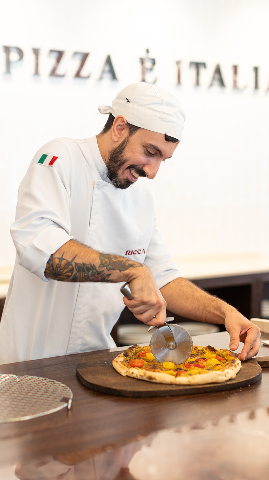 Chef de Ricca Pizzería cortando una pizza artesanal con un cortador profesional en un entorno de cocina italiana.