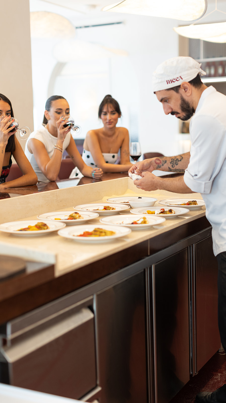 Chef de Ricca Pizzería preparando platos frente a clientes que disfrutan vino en la barra del restaurante.